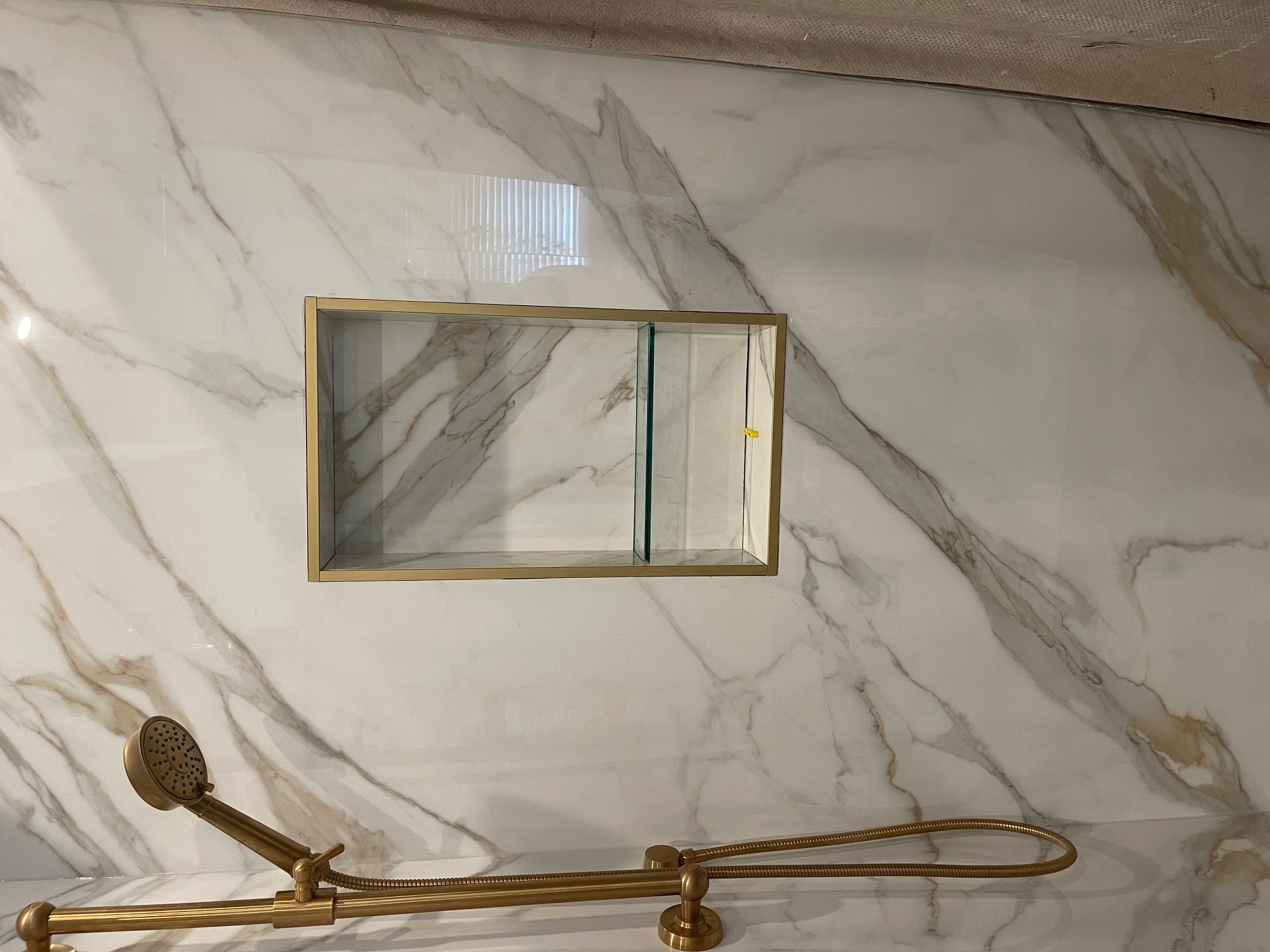 Marble shower wall with brass showerhead, handrail, and recessed glass shelf niche