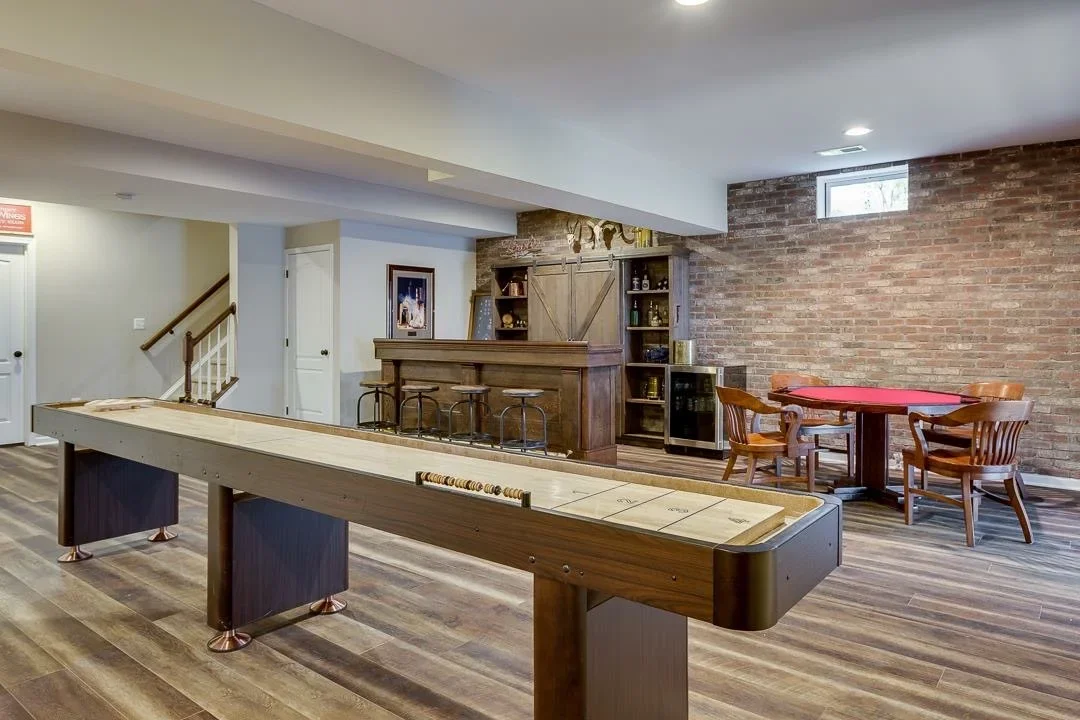 Basement bar with wood cabinetry, brick wall, shuffleboard and billiards tables, recessed lighting