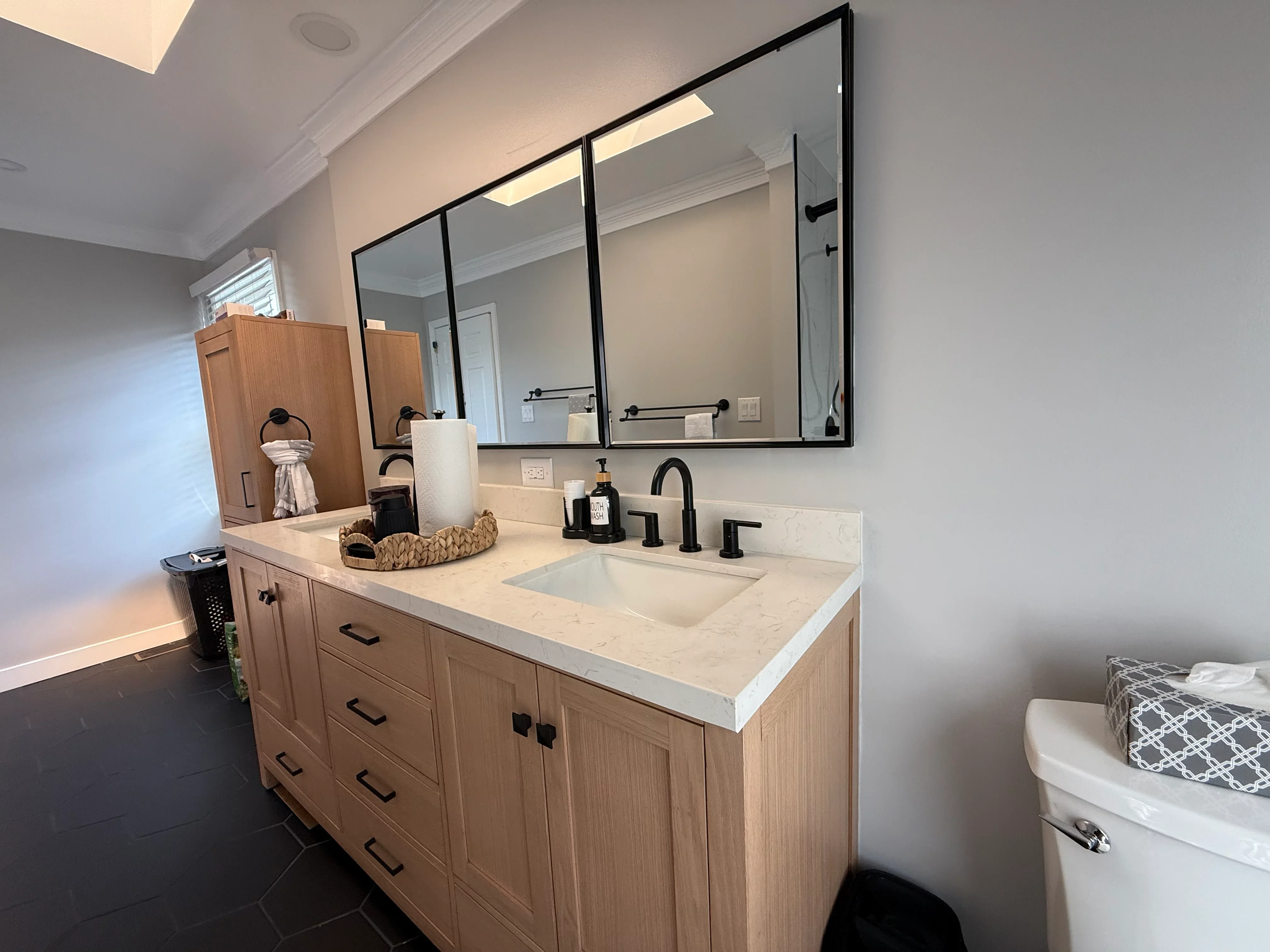 Light oak bathroom vanity with white marble countertop, black-framed mirror, and matte black faucet fixtures